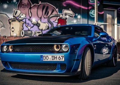 Blauer Dodge Challenger mit schwarzer Motorhaube und deutschen Kennzeichen auf einem Parkplatz.