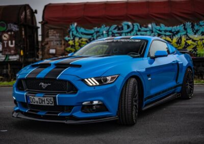 Blauer Ford Mustang mit schwarzen Streifen, frontal aufgenommen, vor einer bunten Wand mit Graffiti.