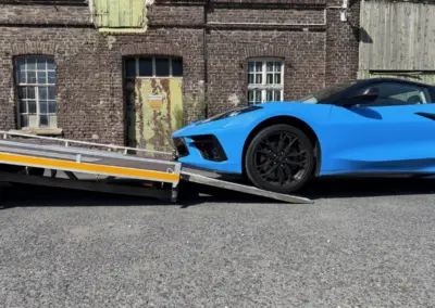 Blauer Sportwagen wird über eine Auffahrrampe auf einem Autotransportanhänger vor einem historischen Backsteingebäude verladen.