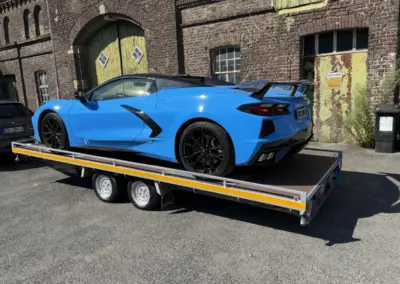 Blauer Chevrolet Corvette C8 auf einem zweiachsigen Autotransportanhänger vor einem historischen Backsteingebäude