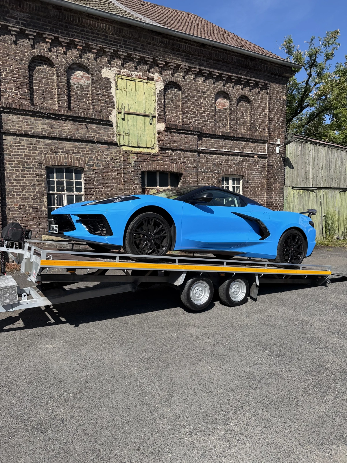 autotransportanhaenger_06 Blauer Sportwagen steht auf einem zweiachsigen Autotransportanhänger vor einem historischen Backsteingebäude in Dortmund.
