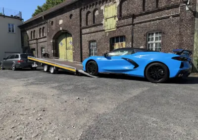Blauer Sportwagen wird auf einem Autotransportanhänger vor einer historischen Backsteinhalle verladen.
